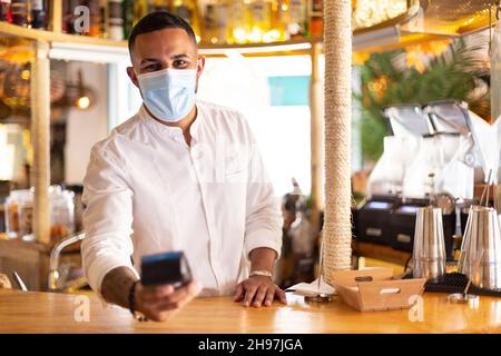 Eleganter lateinamerikanischer Kellner mit einem dataphone in der Hand, um eine Zahlung zu tätigen. Er trägt eine medizinische Maske. Platz für Text. Stockfoto