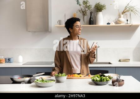 Glückliche tausendjährige verträumte afroamerikanische Frau mit Handy, Kochen. Stockfoto
