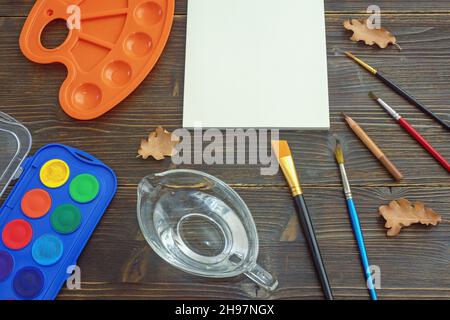 Aquarellmalerei. Skizzenbuch, Palette, Tasse Wasser, Pinsel und Bleistift auf rustikalem Tisch öffnen. Schulkonzept. Freier Speicherplatz für Text oder Bild Stockfoto