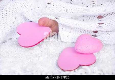 Die Finger des Babys halten ein rosa Herz. Ein Kind in einem weißen Kleid hält mit seinem Fuß ein hölzernes Herz Stockfoto