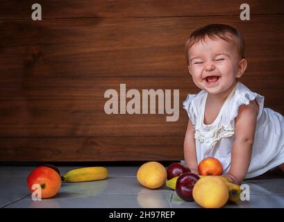 Happy cute Baby Mädchen kriechen auf dem Boden mit Früchten Äpfel, Pflaumen, Bananen, Mango auf einem hölzernen Hintergrund Stockfoto