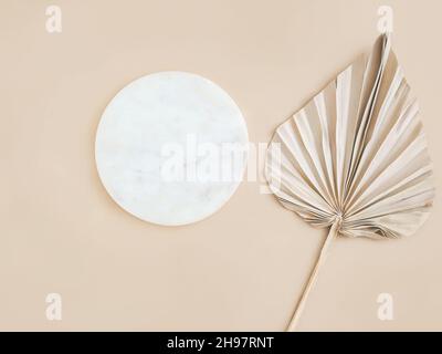 Rundes Podium aus Marmor und trockenes Palmenblatt auf beigem Hintergrund. Ort, Hintergrund für Kosmetik. Draufsicht. Speicherplatz kopieren Stockfoto