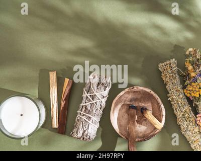 Gegenstände zur spirituellen Reinigung - Salbei und verschiedene Kräuterbündel, palo santo Räucherstäbchen und Kerze auf grünem Hintergrund mit Schatten. Draufsicht. Stockfoto