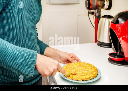 Die Hand eines jungen Mannes hält eine goldene Gabel, um ein frisch zubereitetes spanisches Omelett auf einer weißen Küchentheke zu essen Stockfoto