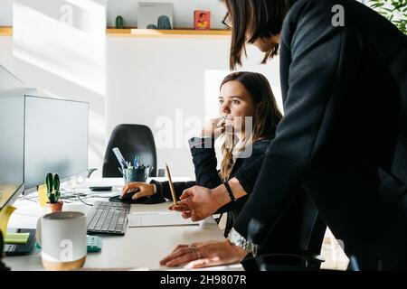 Lächelnde Geschäftsfrau, die einem Kollegen zuhört, vor einem Computer, am Schreibtisch und im Büro. Sie sind in klugen Casuals. Stockfoto