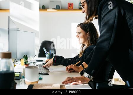 Lächelnde Geschäftsfrau, die einem Kollegen zuhört, vor einem Computer, am Schreibtisch und im Büro. Sie sind in klugen Casuals. Stockfoto