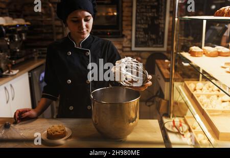 Nahaufnahme einer Köchin, die einen Eierbesen mit Schlagsahne hält. Metallschüsseln mit süßer, köstlicher Creme im Hintergrund. Stockfoto