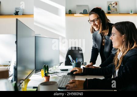 Lächelnde Geschäftsfrau, die einem Kollegen zuhört, vor einem Computer, am Schreibtisch und im Büro. Sie sind in klugen Casuals. Stockfoto