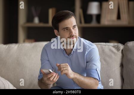 Nachdenklicher, positiver junger Mann, der Mobiltelefon benutzt und auf der Couch sitzt Stockfoto