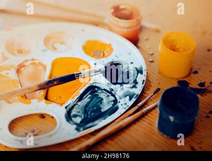 Auf dem Holztisch ist ein kreatives Chaos zu sehen. Die Palette ist mit Gouache-Farben gefärbt, daneben liegen Pinsel und Gläser mit Farben Stockfoto