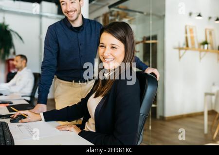 Lächelnde Geschäftsfrau, die einem Kollegen zuhört, vor einem Computer, am Schreibtisch und im Büro. Sie sind in klugen Casuals. Stockfoto