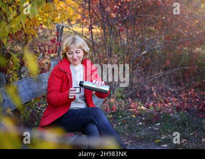 Eine angenehme Frau von 50-60 Jahren in einem Herbstpark, die auf einer Holzbank sitzt, gießt Tee aus einer Thermoskanne, um sich warm zu halten. Stockfoto