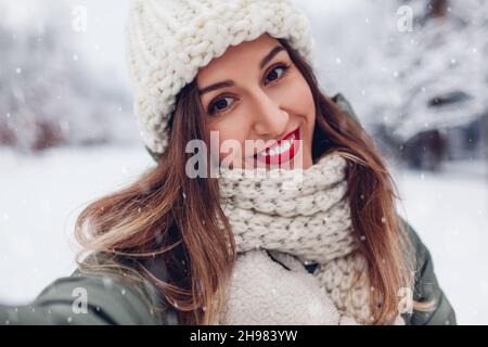 Glückliche junge Frau macht Selfie im verschneiten Winterpark in warmen Strickkleidung und rotem festlichem Lippenstift bei schönem Wetter. Nahaufnahme im Hochformat Stockfoto