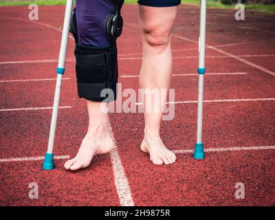 Sportlerin mit Krücken, Handgelenkstütze und Kniestütze, gebändertes Bein. Frau läuft auf roter Lauffläche Stockfoto