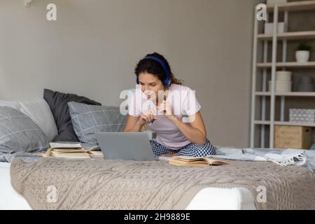 Nachdenkliche Frau in kabellosen Kopfhörern, die online studiert und auf dem Bett sitzt Stockfoto