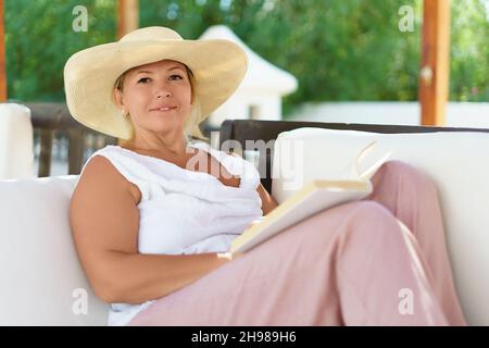 Eine lächelnde kaukasische Frau mittleren Alters las an einem sonnigen Sommertag im Garten ein Buch auf dem Sofa. Stockfoto