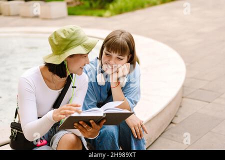 Multirassische zwei Frauen, die ein Buch lesen und lächeln, während sie sich im grünen Park ausruhen Stockfoto