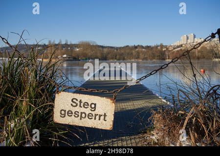 Schild an einer rostigen Kette mit der Aufschrift: Dock gesperrt (steg gesperrt). Schwarze Buchstaben auf weißem Brett. Gefrorener blauer See und Wolkenkratzer im Hintergrund, Stockfoto