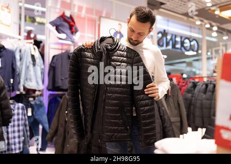 Der unrasierte, gutaussehende Mann mit Brunet wählt in einem Bekleidungsgeschäft in einem Einkaufszentrum eine warme schwarze Jacke. Oberbekleidung und Einkaufen Stockfoto