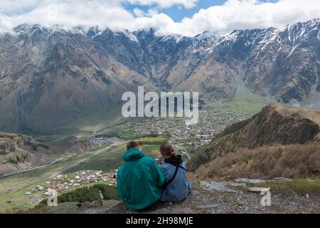 Junges Paar mit Blick auf Stepantsminda in seinem Tal von der Gergeti Trinity Church. Region Mzcheta-Mtianeti, Georgien, Kaukasus Stockfoto