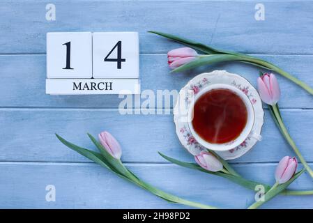 Dampfende heiße Tasse Tee und rosafarbene Frühlingsblumen über einem rustikalen blauen Holztisch, aufgenommen von oben. Kalenderblöcke für März 14th. Stockfoto