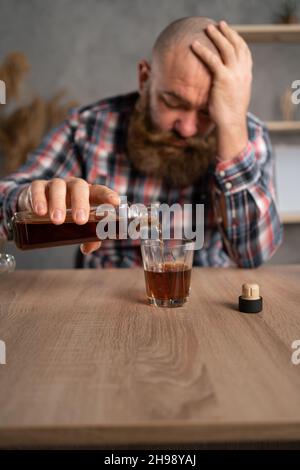 Depressionen und Alkoholismus bei Männern. Ein erwachsener bärtiger Kerl sitzt selbst mit Alkoholflaschen an einem Tisch. Er hält Brandy in den Händen und gießt ihn hinein Stockfoto