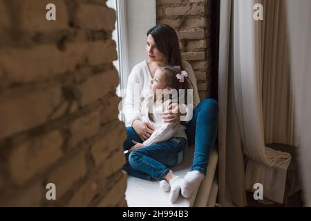 Eine junge, schöne Mutter sitzt auf einer hölzernen Fensterbank in einem weißen Pullover und Jeans mit den Armen um ihre junge Tochter. Stockfoto