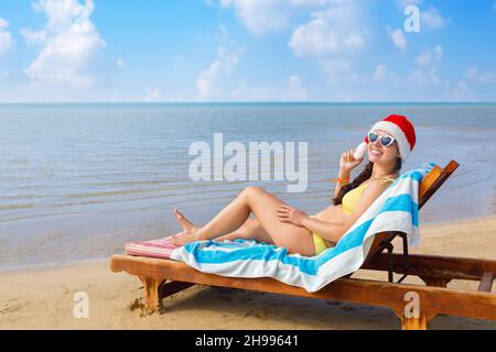 Fröhliche junge Frau mit Weihnachtsmütze und Sonnenbrille auf der Sonnenliege Stockfoto