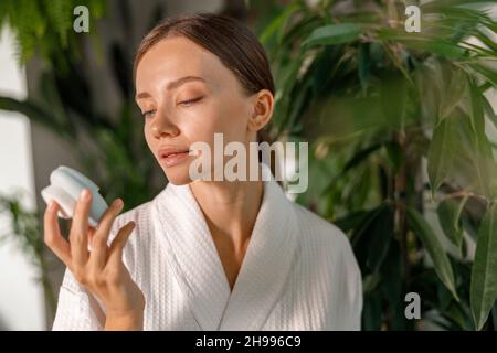 Junge Frau im Bademantel versucht, Silikon-Gesichtsbürste für die Haut Peeling verwenden, mit Schönheitsroutine im Badezimmer mit Pflanzen dekoriert Stockfoto