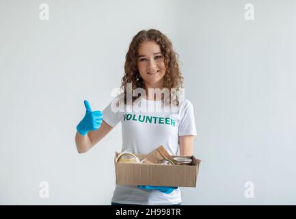 Aufgeregter Aktivist hält eine freiwillige Schachtel mit Spenden und zeigt den Daumen nach oben, lächelt die Kamera über hellem Hintergrund an Stockfoto