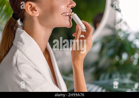 Seitenansicht einer lächelnden Frau im Bademantel, die Lippenbalsam aufbringt, sich um ihre Lippen kümmert und im Badezimmer steht Stockfoto