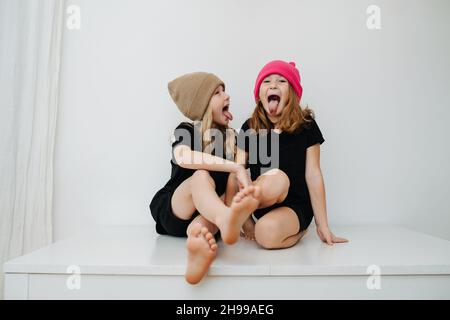 Dumme, verspielte Geschwister in bunten Strickmützen sitzen auf einem Tisch und halten ihre Zungen heraus Stockfoto