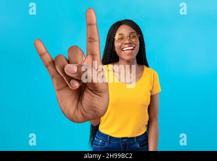 Fröhliche afroamerikanische Frau mit großer Rock-Geste, lächelnd auf die Kamera, Sonnenbrille auf blauem Hintergrund Stockfoto