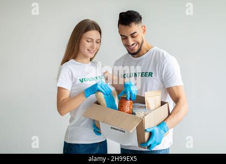 Fröhliche Aktivisten verpacken Spenden von Lebensmitteln, legen Dosen und Gläser in einen Karton und stehen über einer hellen Wand Stockfoto