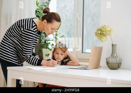 Mama hilft kleinen Mädchen zu zeichnen. Sie steht über dem Tisch und zeichnet mit einem Stift. Das Mädchen schaut genau zu und lehnt sich an ihre Hand. Stockfoto