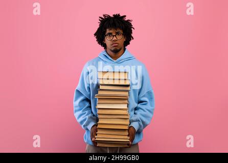 Unglücklicher schwarzer Teenager, der riesige Bücherstapel hält und mit gelangweiltem Gesichtsausdruck auf rosa Hintergrund auf die Kamera schaut Stockfoto
