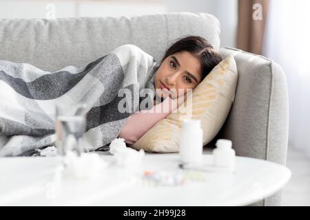 Frustration junge hindu-Dame mit Karli bedeckt, an Grippe leiden, liegt auf dem Sofa im Zimmer mit Wasser und Pillen Stockfoto