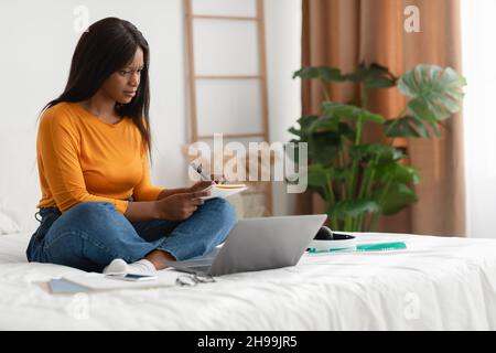 Besetzt Schwarz Weiblich Nehmen Notizen Lernen Mit Laptop Im Schlafzimmer Stockfoto