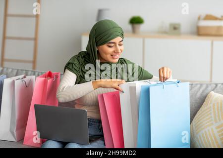 Junge arabische Frau im Hijab sitzt auf dem Sofa mit Laptop, schaut in Geschenktüten, überprüft die gelieferte Online-Bestellung zu Hause Stockfoto