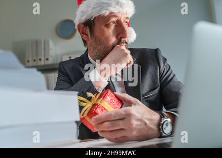 Besorgter Geschäftsmann mit Weihnachtsmannhut, der während der Weihnachtsfeiertage aus dem Bürofenster schaut, selektiver Fokus Stockfoto