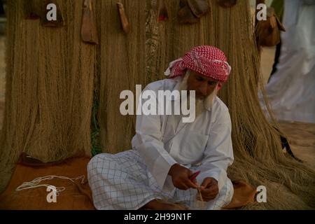 Ein traditioneller arabischer Handwerker webt Fischnetz im Katara Kulturdorf, Doha Katar Tageslichtansicht mit Daus im arabischen Golf im Hintergrund Stockfoto