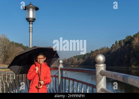 Porträt eines Teenagers mit einem Regenschirm auf dem Hintergrund des Flusses und des Herbstwaldes Stockfoto