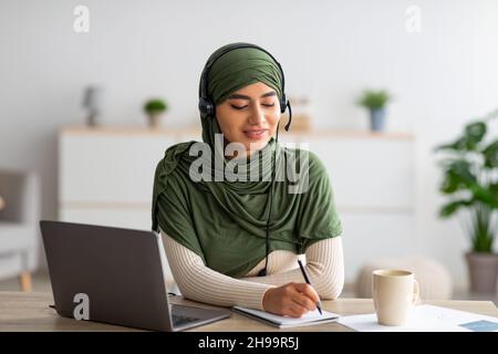 Junge arabische Frau im Hijab, die am Laptop arbeitet, Kopfhörer trägt und sich während des Webinars zu Hause Notizen macht Stockfoto