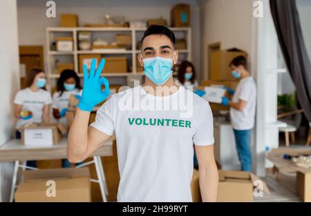 Konzept der Freiwilligenarbeit. Junger arabischer Mann mit medizinischer Maske und Handschuhen, der eine gute Geste zeigte und im Charity Center arbeitete Stockfoto