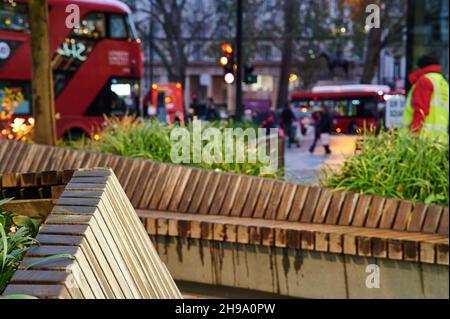 London Dämmerung Straßenszene mit verschwommenem Hintergrund mit london Bus, Fußgänger, Pflanzen und Holzsitze. Keine Marken sichtbar. Stockfoto