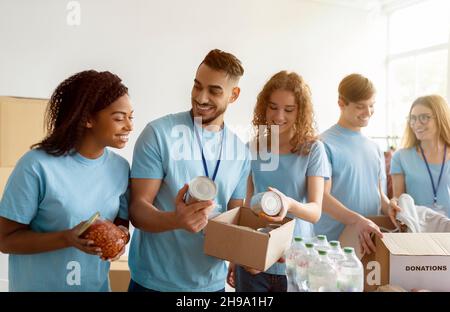 Konzept der humanitären Hilfe. Junge, vielfältige Freiwillige sortieren und verpacken Konserven in Kartons und arbeiten zusammen Stockfoto