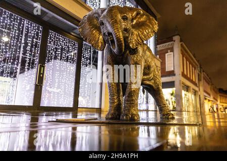 London, Großbritannien. 5th Dez 2021. Die Menschen gehen im Regen an einer Skulptur des Elefanten Mukkoka vorbei, wobei die Lichterdekoration eines nahegelegenen Gebäudes die frühe Abendszene erhellt. Mukkoka, der Elefantenbaby, ist Teil der „Herde der Hoffnung“, einer 21-köpfigen Herde von Bronzeelefanten, die von den Künstlern Gillie und Marc geschaffen wurde und nun Spitalfelder durchstreift, um das Bewusstsein für den Schutz und gefährdete Arten zu schärfen. Kredit: Imageplotter/Alamy Live Nachrichten Stockfoto