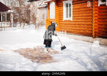 Frau putzt Teppich. Kaukasische ältere Frau ist in der traditionellen Methode der ökologischen Teppichreinigung mit Neuschnee und Besen im Hinterhof auf engagiert Stockfoto