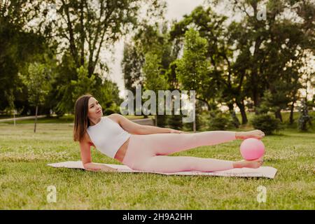 Friedliche, lächelnde Frau in pinkfarbenen Kleidern und einem kleinen Gummiball, die im Sommer bei Sonnenuntergang im Park Pilates auf rosa Matten macht Stockfoto