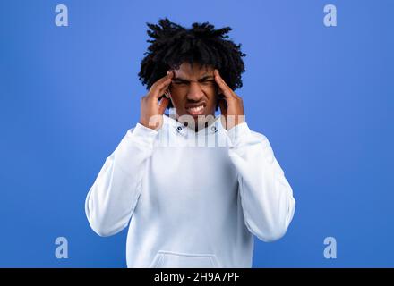 Afroamerikanischer Teenager, der unter starken Kopfschmerzen leidet und Tempel über blauem Studiohintergrund massiert Stockfoto